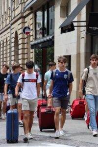 Casual young men walk down a city street with suitcases, showcasing travel style.